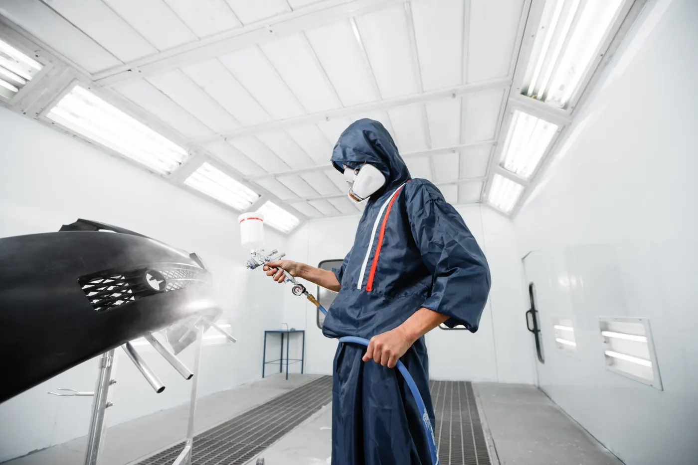 A painter spraying a bumper panel inside a spray booth cabin (general workshop scene)